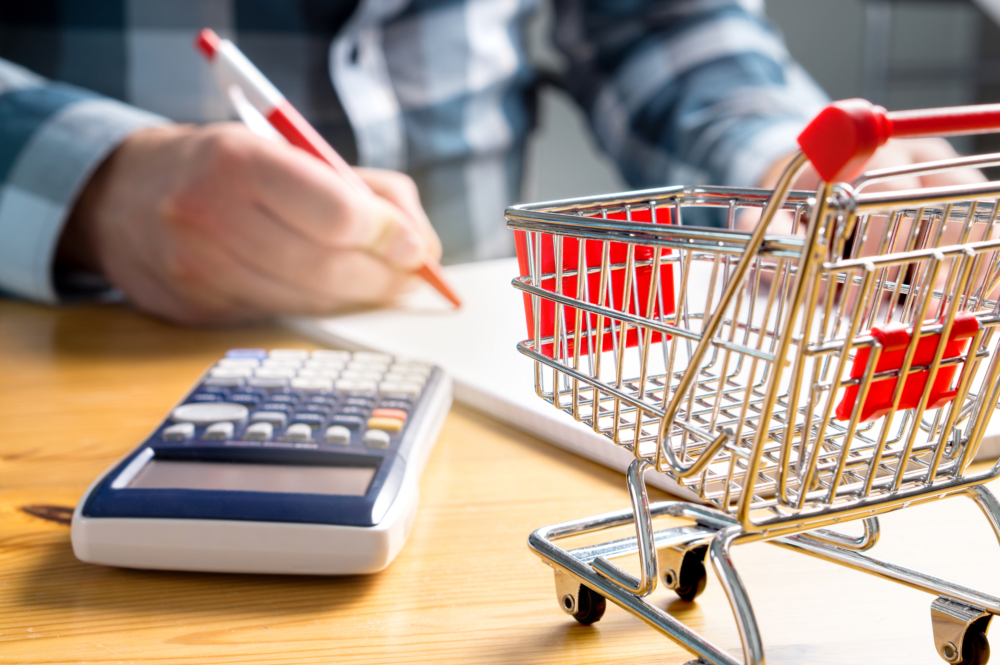 Prices are rising, but by how much? Hand holding a pen on a notepad, with a calculator and a mini shopping cart on a table.
